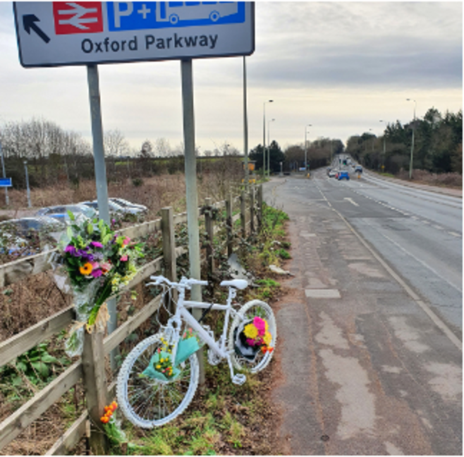 A 'ghost bike' adorned with flowers, parked on the side of a main road near a train station.  A bicycle roadside memorial.
