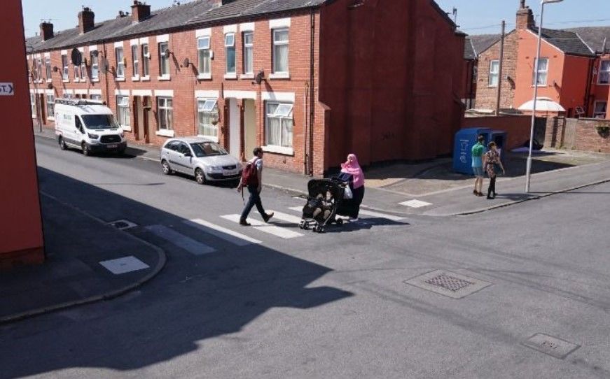 A man and a woman pushing a buggy walk over a zebra crossing across a side road on a small residential street