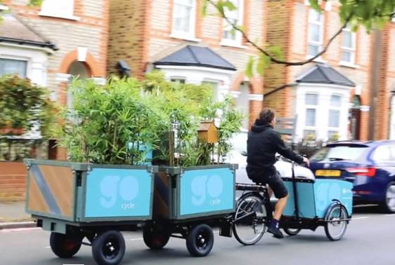 A cargo-bike rider towing two trailers containing plants and a bird box