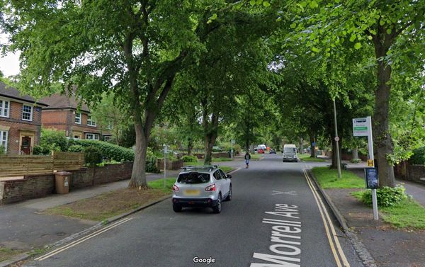 A residential street with bus stops and lined by trees being used by a scooter rider and cars